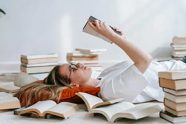 Young woman with glasses reading a book while lying on the floor surrounded by open books