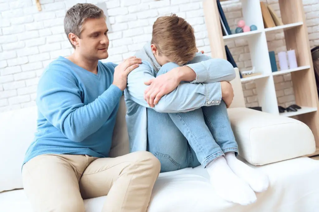 Father comforting his teenage son who is sitting on a couch, appearing upset