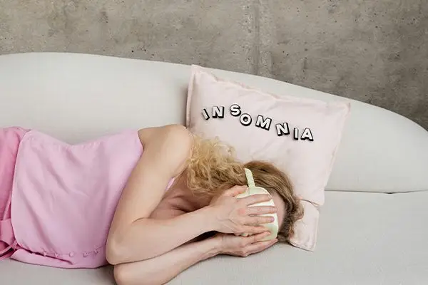 Woman lying on a couch covering her face, with a pillow labeled “Insomnia”