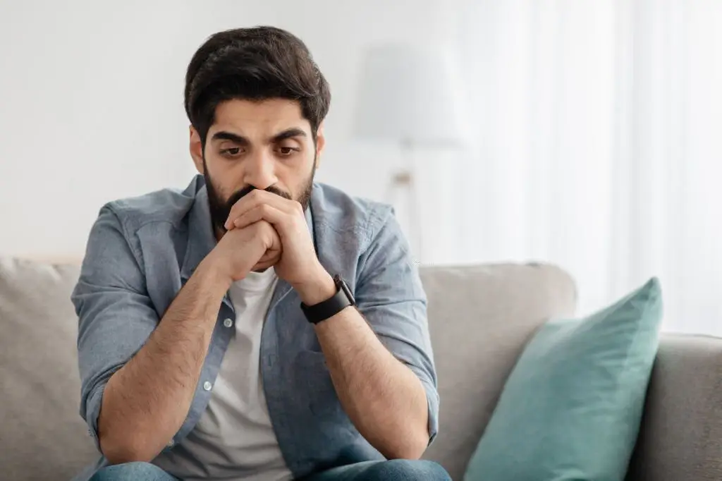 Anxious man sitting on a couch with hands clasped near his mouth, looking worried